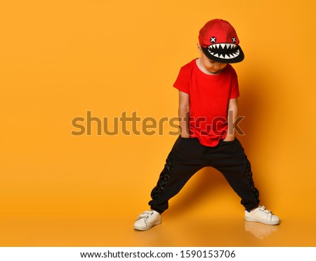 Little cute 4 year old boy in wide dark pants with pockets and a red T-shirt. A merry cap with shark teeth. Standing on a yellow background, put his hands in his pockets and looks down