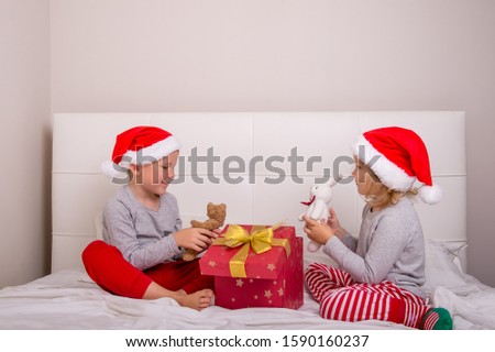 Funny boy and girl having fun in bedroom on Christmas morning. Kids in Santa Clause red hats open gift box. Happy xmas and New Year. Children playing in bed.