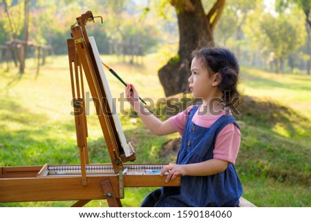A little girl is sitting on the wooden bench and painted on the canvas placed on a drawing stand, Be a part of learning outside of the school in the nature park