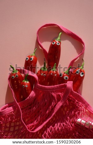 Sweet Small Red Peppers In The Pink Cotton Mesh Bag. Funny faces with wobbly eyes. Fire Red And Soft Pink Colors Background.