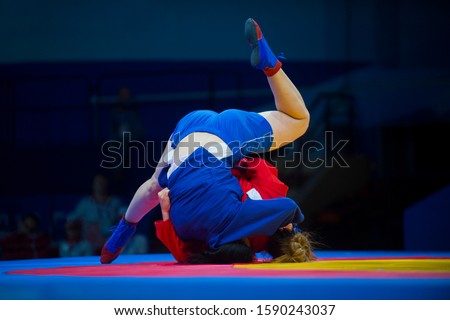 Two woman in blue and red wrestling on a yellow wrestling carpet in the gym

