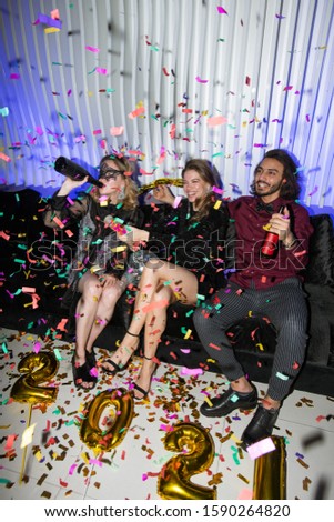 Young joyful friends with beer sitting on soft couch while having fun during new year party in the night club