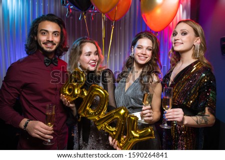 Three cheerful girls and young elegant man holding flutes of champagne and balloons while enjoying birthday party