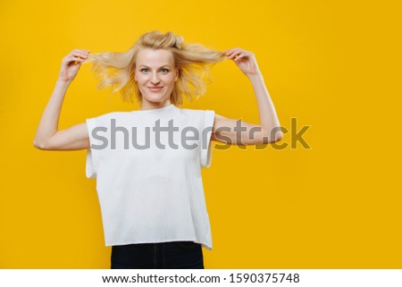 Playful grimacing blonde woman in a white sleeveless shirt is fooling around, making funny faces over yellow background. She's pulling her hair to sides.