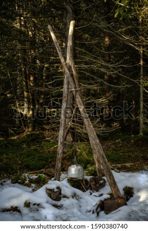 A vertical picture of metallic pot hanging from woods surrounded by greenery and snow