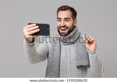 Funny man in gray sweater, scarf isolated on grey background. Healthy lifestyle, ill sick disease treatment, cold season concept. Hold thermometer doing selfie shot on mobile phone, making video call