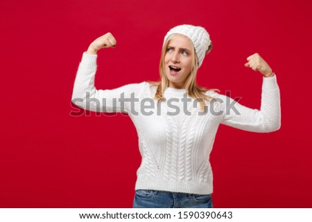 Strong young woman in white sweater, hat isolated on red wall background, studio portrait. Healthy fashion lifestyle, people emotions, cold season concept. Mock up copy space. Showing biceps, muscles