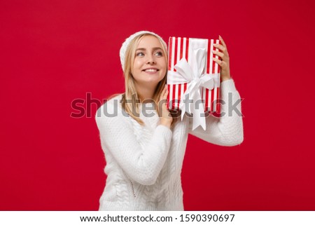 Pretty young woman in white sweater, hat isolated on red wall background. New Year 2020, Valentine's Day, birthday, holiday concept. Mock up copy space. Hold striped present box with gift ribbon bow