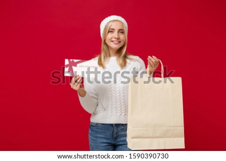 Smiling young woman in sweater, hat isolated on red background. Healthy fashion lifestyle, cold season concept. Mock up copy space. Hold package bag with purchases after shopping, gift certificate