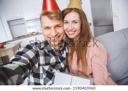 Young man and woman family sitting together on the coach wearing birthday caps for celebration taking selfie photos smiling close-up