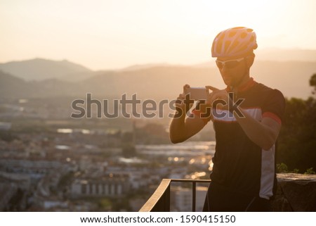 Cyclist Taking Picture With Smart Phone. Traveller Make Photo On Smartphone, Using Phone In Hand, Travel Blogger. Sportive man with bicycle use phone takes a photo in spain on a mountain at sunset.