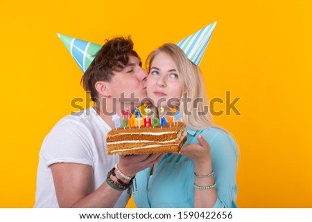 Funny young couple in paper caps and with a cake make a foolish face and wish happy birthday while standing against a yellow background. Concept of congratulations and fooling around.