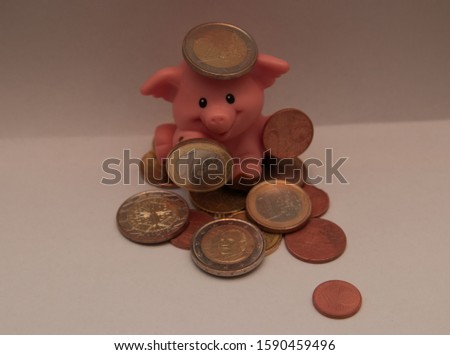 Small pink pig smiling, sitting on euros coins and holding some money. Balancing money on its head and holding some euros in it hands