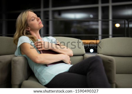 Young blonde woman dreaming and relaxing while holding a book, sitting on an armchair, legs crossed 