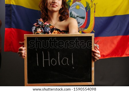 Portrait of young Latin American woman student holding a chalkboard with Hello word on her native language before the Ecuadorian flag..
