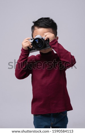 Portrait of little boy wearing red sweater standing isolated on grey background taking photo with film camera. 
