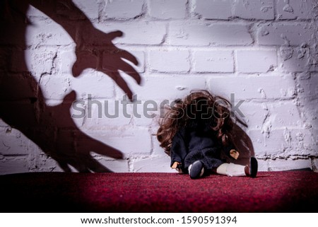 A doll sits on the floor and leans against a wall. The shadows of hands reach for her. Concept: violence and abuse