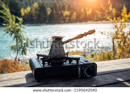 Copper turk with coffee on gas burner on the background of river and hills. Coffee preparing on the banks of mountain river on portable gas stove during hiking trip. Tourism concept.
