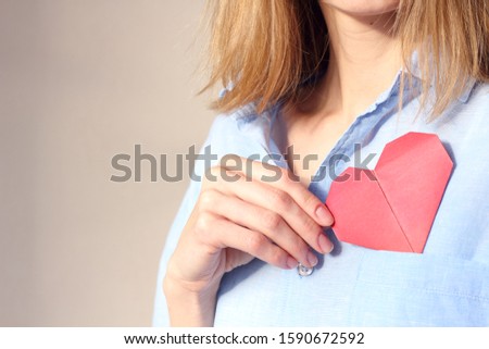 cropped image of woman holding origami red paper heart, puts valentine in breast pocket. Insurance, charity donation concept. Place for text, copy space, selective focus