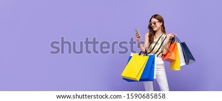 Trendy beautiful young Asian woman carrying colorful bags shopping online with mobile phone isolated on purple banner background with copy space Royalty-Free Stock Photo #1590685858