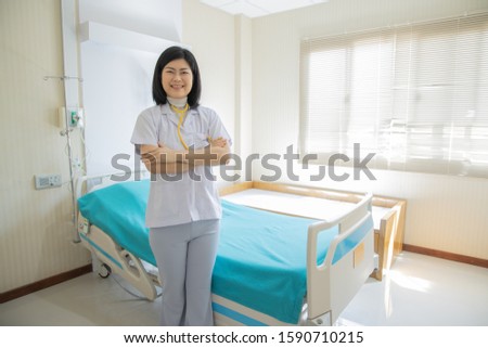Asian female doctor (nurse or specialist) stood smiling at the patient's bed. Providing kindness, warmth, trust in healing concept. (Wearing a white uniform and stethoscope). Look camera. Cross arm.