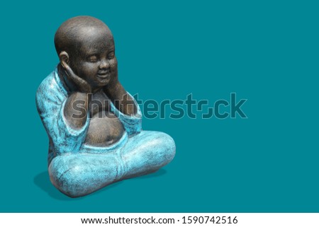 Little buddha monk covers his ears with his hands as a symbol - not to hear evil. Religious sculpture on a turquoise background.