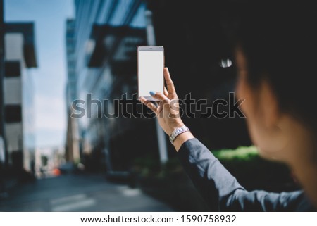 Back view of young female dressed in black jacket taking picture of city with mobile phone in sunny day in downtown