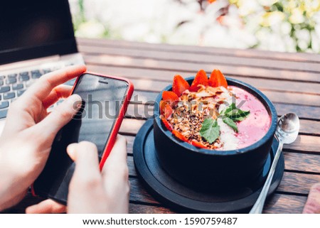 Faceless from above view of person using mobile phone with touch screen enjoying colorful exotic breakfast bowl while sitting in cafe with laptop 