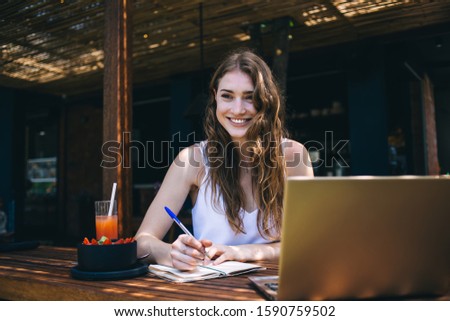 Young pretty female author with bright smile sitting in cafe in morning and writing down ideas in notebook on dark blurred background Royalty-Free Stock Photo #1590759502