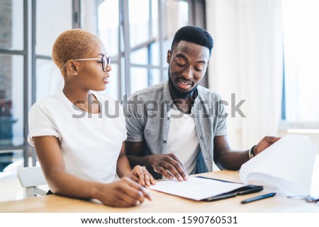 Focused black couple sitting at table in apartment in front of realtor reading estate contract with attention and talking with concentration