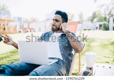Smart relaxed smiling young ethnic male in casual denim shirt listening to music with laptop and earphones spending time in sunny park