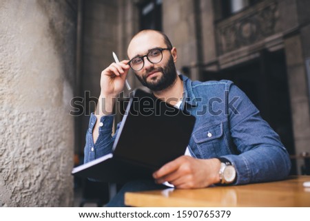 Thinking adult writer sitting in pavement cafe at table and holding black notebook and pen while setting glasses right and looking away