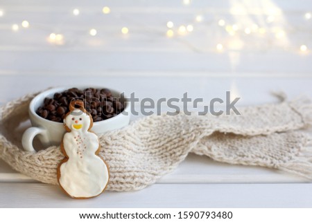 a white cup with roasted coffee beans and a cheerful snowman is wrapped in a knitted scarf on the table. winter festive morning coffee