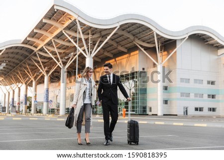 Photo of smiling colleagues man and woman in formal wear walking with suitcase on parking near airport outdoors