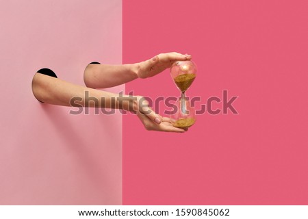 Vintage sandglass in a female's hands from two holes in the wall on a duotone pink background, copy space. Royalty-Free Stock Photo #1590845062