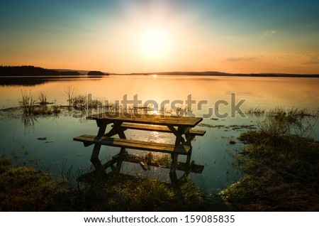 beautiful picture of a table with bench by a lake with the sun and sunbeams in the sky