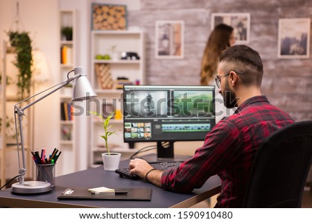 Back view of creative filmmaker working on a movie on laptop. Girlfriend in the background. Royalty-Free Stock Photo #1590910480