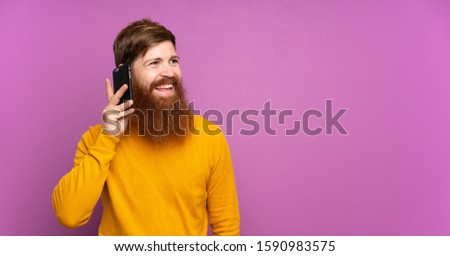 Redhead man with long beard over isolated purple background keeping a conversation with the mobile phone