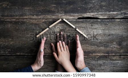 Conceptual image of family and adoption - mum and dad forming a house shape with their hands to shelter their biological and adopted child. Royalty-Free Stock Photo #1590997348