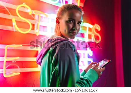 Image of beautiful young caucasian woman holding smartphone over multicolored neon text sign