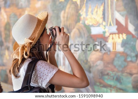 Back side traveling Asian woman in white dress under straw hat shooting camera to beautiful wall painting at Wat phra kaew, on of the most sacred Buddhist temple in Thailand.  Travel and vacation.