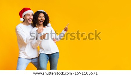 Happy interracial spouses in santa hats pointing with fingers at copy space on yellow background, demonstrating place for your ad, panorama