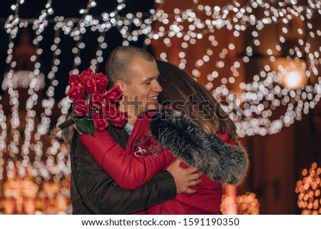Couple. Love. Holiday. Outdoors. Woman is holding a bouquet of roses and smiling while hugging her man at winter night in the city street