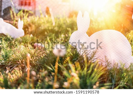 The concept of Easter holidays. Decorative handicraft handmade product in the form of a lying white rabbit, located next to the painted eggs. In the background the grass and the house. Sunlight