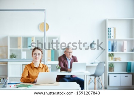 Portrait of young businesswoman in eyeglasses working on laptop and looking at camera with mature man working in the background