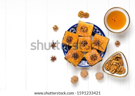 Traditional arabic dessert baklava with cashew, walnuts, raisins on plate, bowl of tea with Uzbek national ornament on white wooden table. Homemade baklava with nuts and honey Top view Flat Lay
