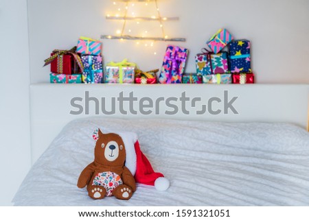 Cozy toy bear in santa hat on the bed on background of colourful gift boxes and Christmas tree made of wooden sticks with garland on the wall. Simple, minimal interior design and decor of child bedroo