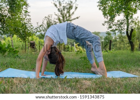 Side view portrait of the young girl in white undershirt doing yoga exercise on the blue mat among the garden, standing in varius poses, outdoor photo