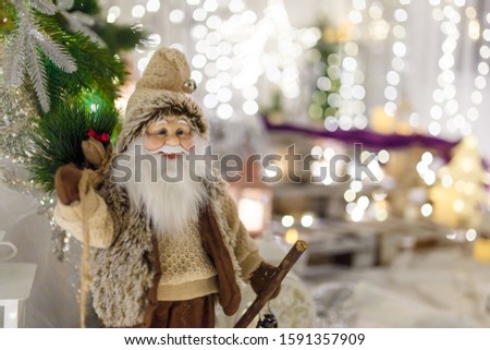 Santa Claus toy in brown clothes. Fir tree branches and Christmas decorations on blurry background with warm garland lights and bokeh. Winter holidays concept. 