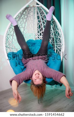 Ugly aged woman in a sweater in a white wicker hanging chair in the room with curtains. Old model during photoshoot in studio